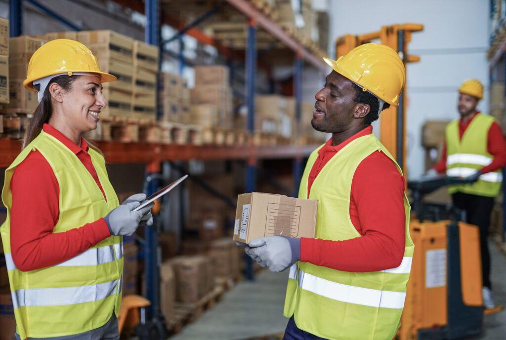 Staff Working In Warehouse With Protective Wear Young African Man With Package And Mature Latin Woman With Digital Tablet